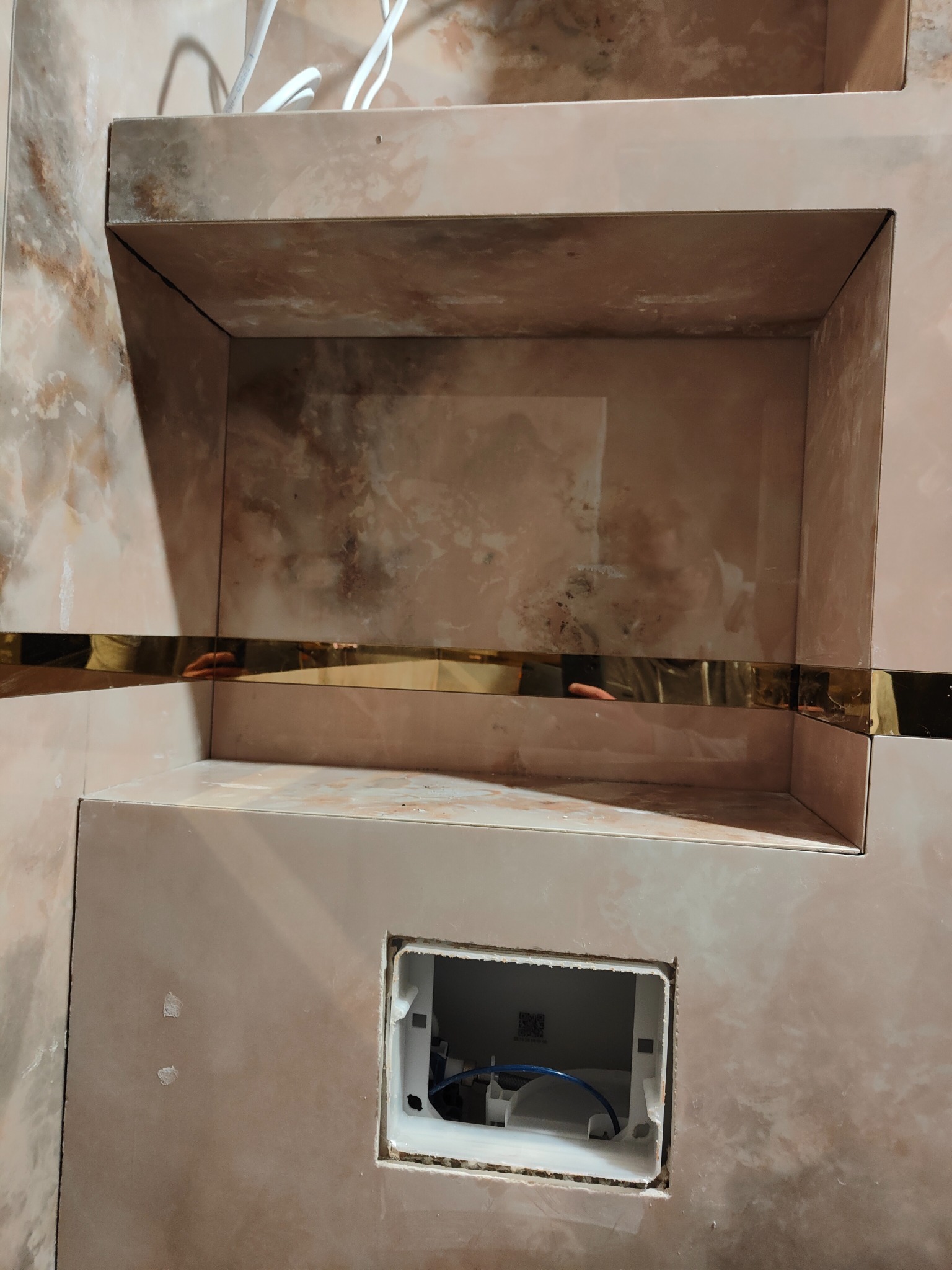 Niedokończona wnęka z półkami w łazience, wyłożona płytkami imitującymi marmur, z widocznym fragmentem instalacji podtynkowych.