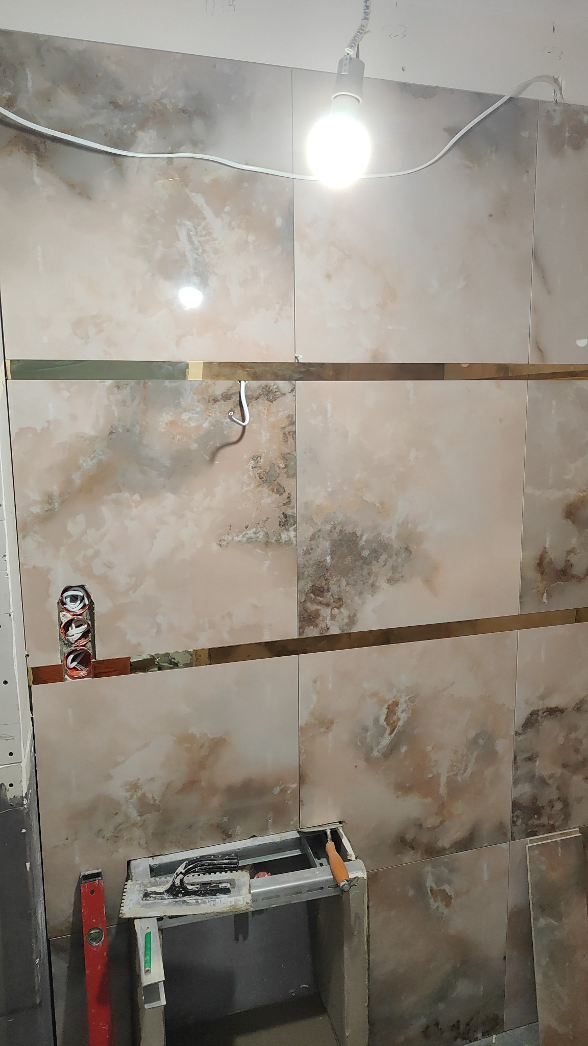 Układanie płytek imitujących marmur w łazience, widoczna poziomica, przecinarka do glazury i puszka elektryczna z przewodami.