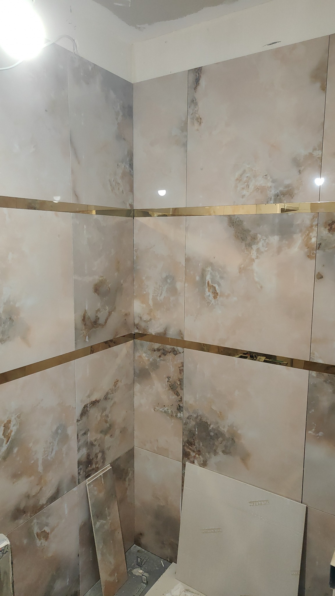 Ściana łazienki wyłożona płytkami imitującymi marmur z złotymi listwami dekoracyjnymi, widoczne niedokończone prace i płyty na podłodze.