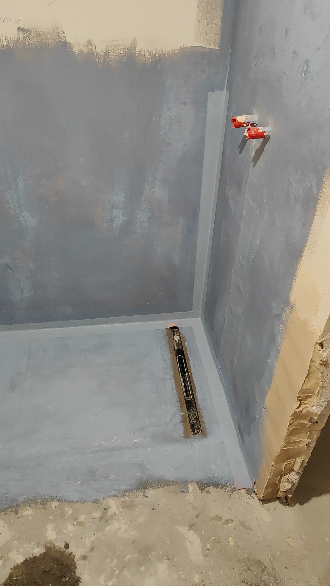 Przygotowana do hydroizolacji strefa prysznicowa z widocznymi wystającymi rurami, poziomica na podłodze i częściowo pomalowane ściany na szaro.