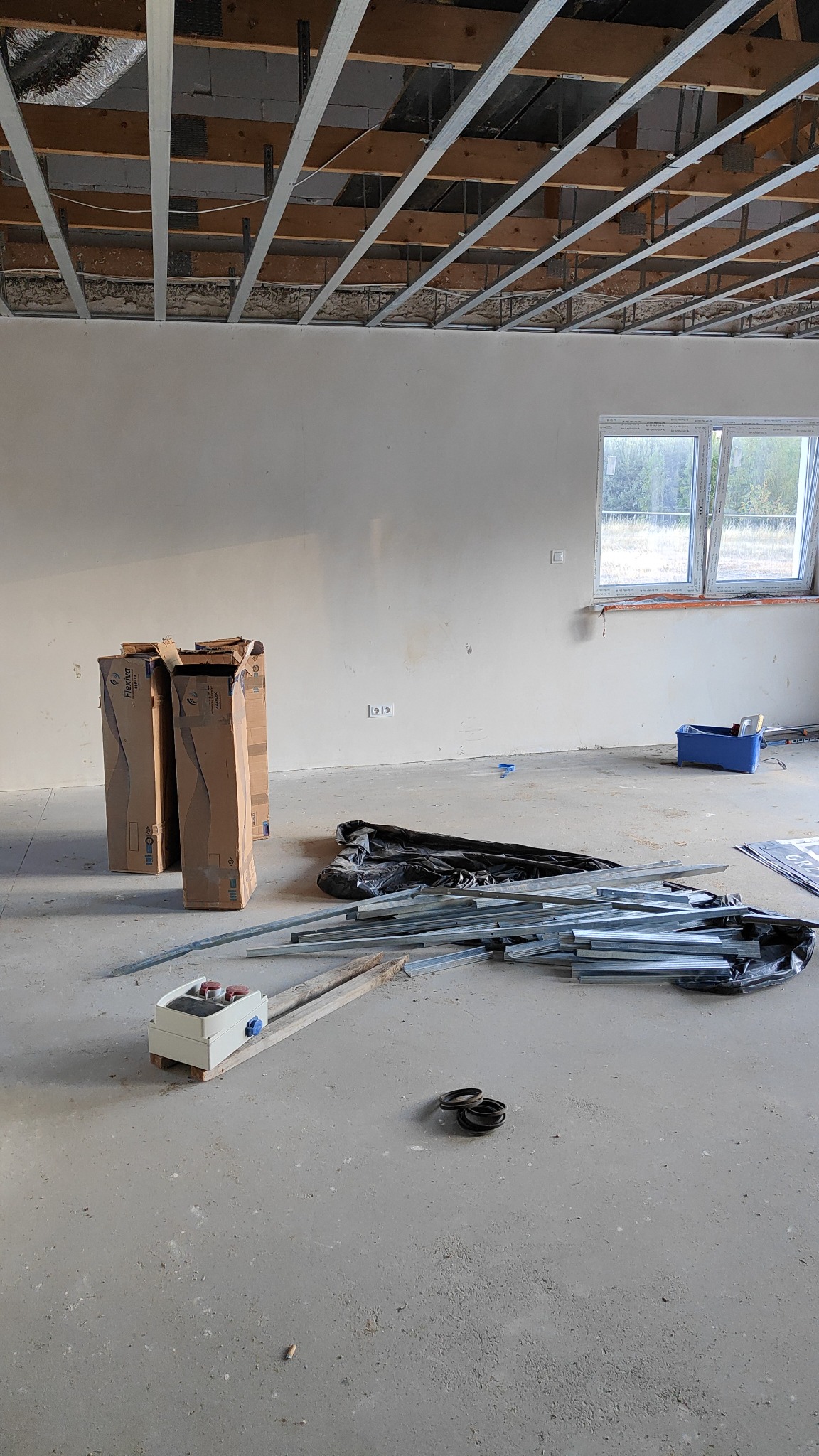 Wnętrze w trakcie prac wykończeniowych: widoczne metalowe profile sufitowe, kartony z materiałami budowlanymi, narzędzia i akcesoria na betonowej podłodze, ściany przygotowane do malowania, okno...