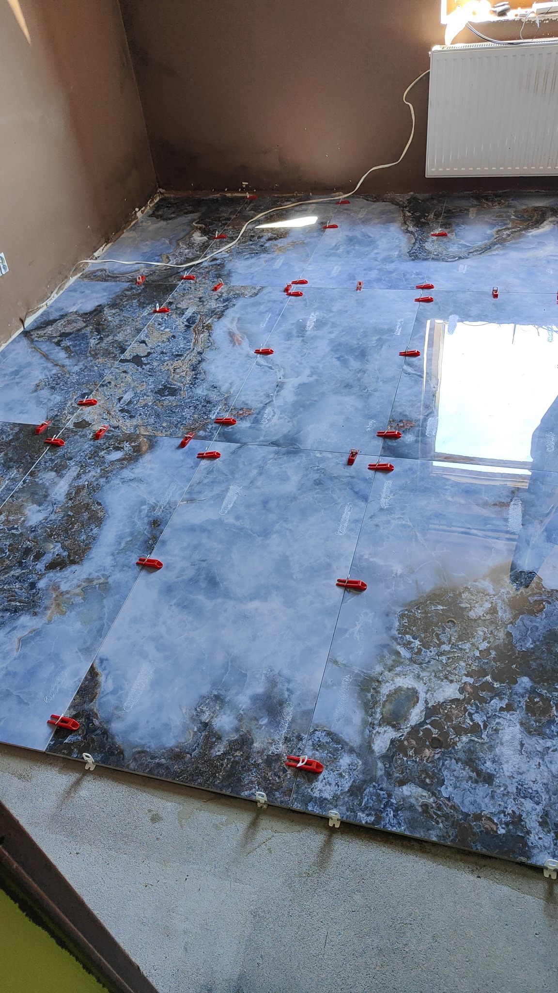 Układanie płytek podłogowych o wzorze imitującym marmur, z użyciem czerwonych i białych klipsów poziomujących, widoczne niedokończone krawędzie pod ścianą.