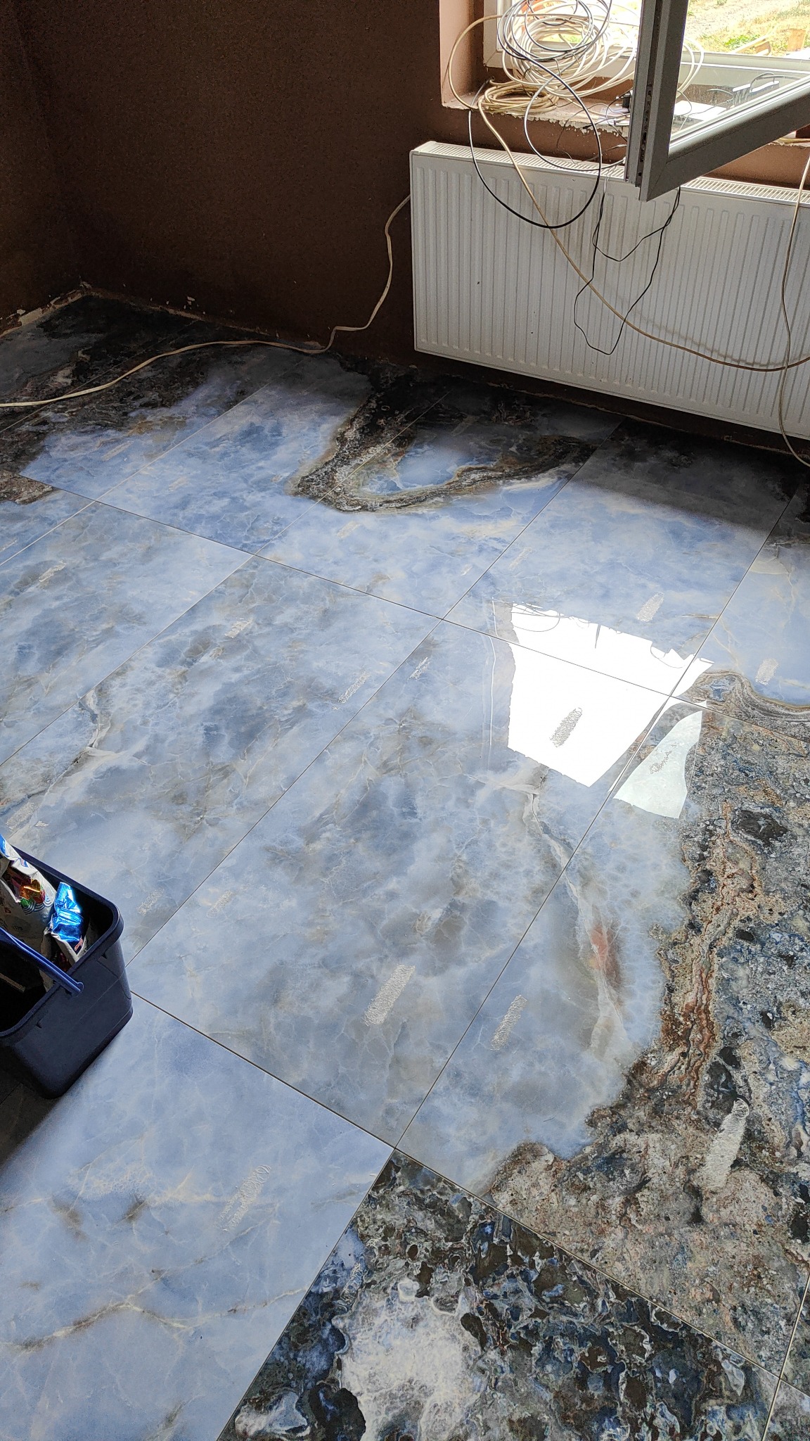 Ułożone, błyszczące, marmurkowe płytki podłogowe w odcieniach błękitu i szarości, widoczny fragment ściany, grzejnik i okno z kablami, w rogu wiaderko z narzędziami.