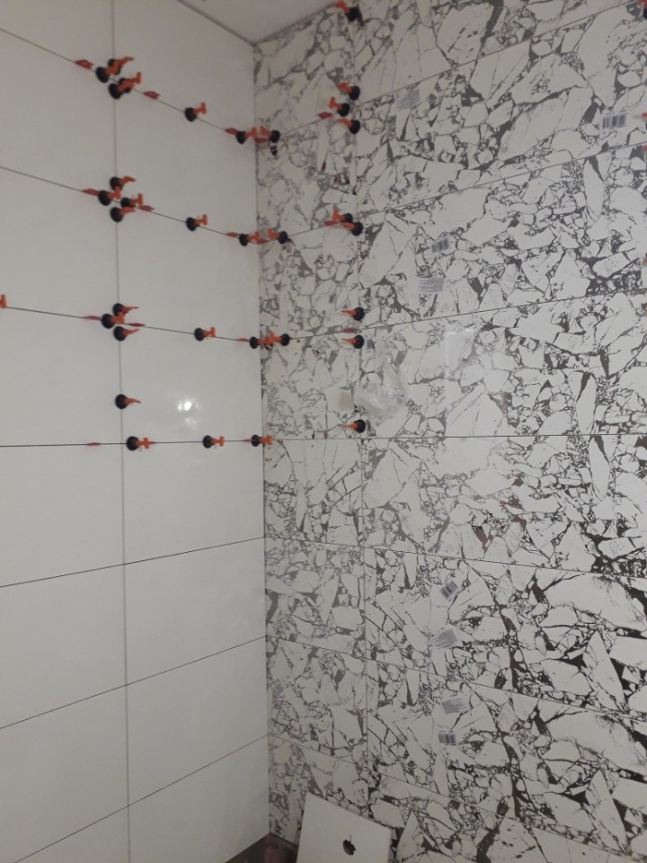 Układanie płytek w łazience: białe, gładkie płytki na jednej ścianie kontrastują z płytkami imitującymi marmur na drugiej, z użyciem poziomujących klipsów i krzyżyków.