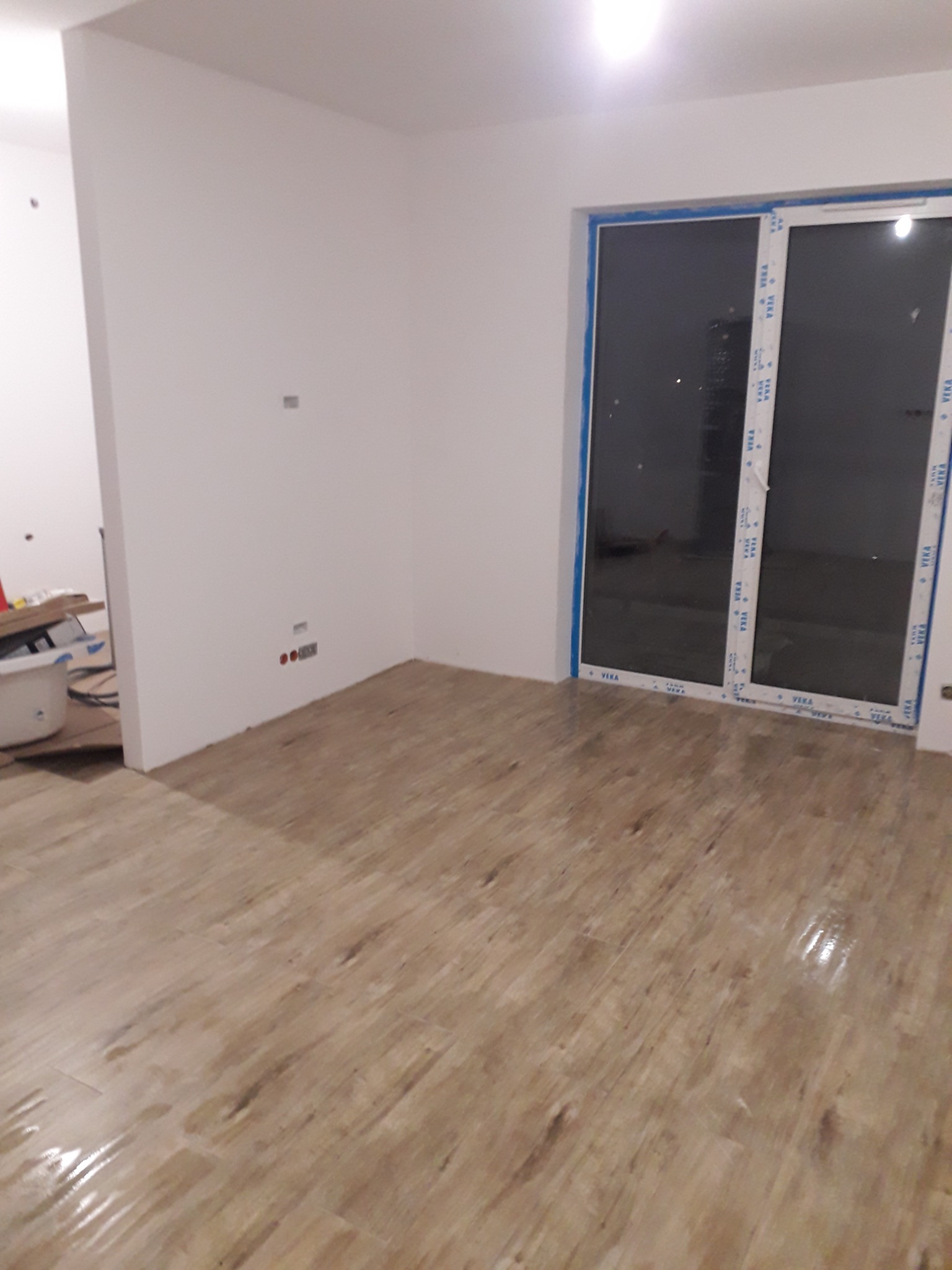 Nowe panele podłogowe w jasnym odcieniu drewna, widoczne gniazdka elektryczne na świeżo pomalowanej białej ścianie, duże okno przesuwne z niebieską folią ochronną na ramie.