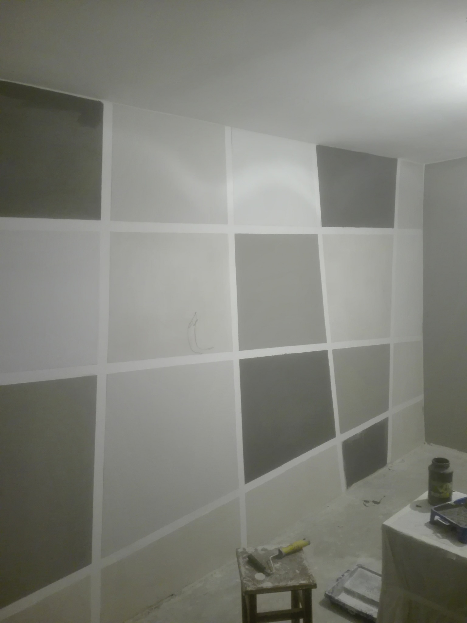 Ściana w trakcie malowania z geometrycznym wzorem tworzonym za pomocą taśmy malarskiej, widoczne narzędzia malarskie i farby.