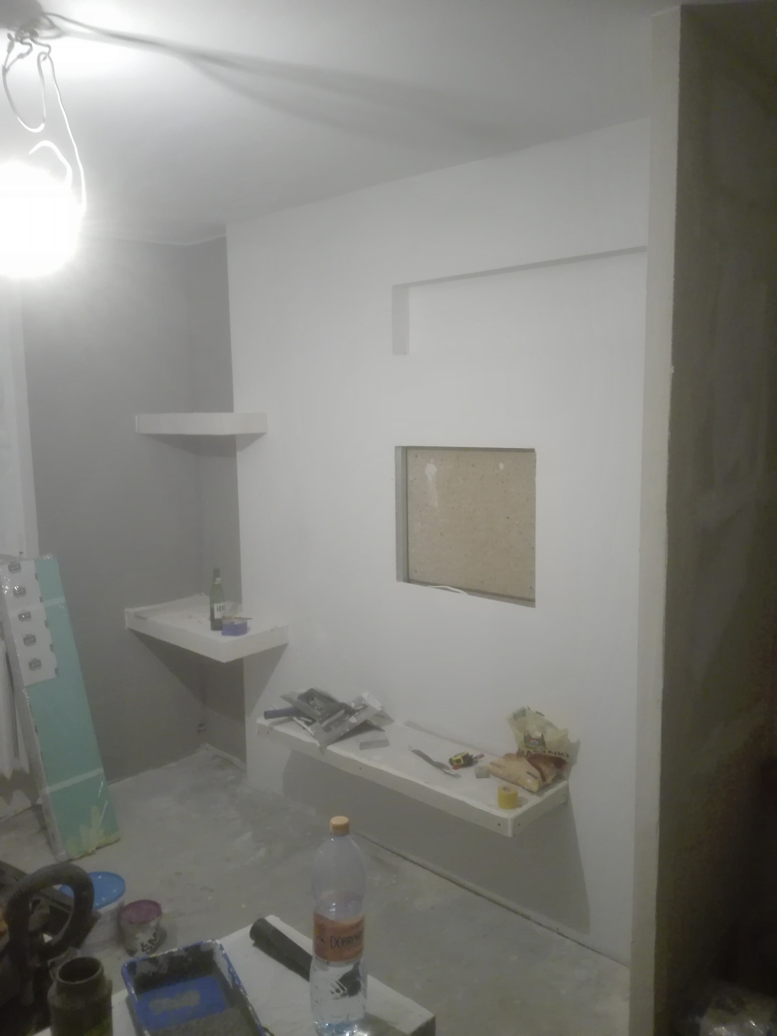 Wnętrze pokoju w trakcie prac wykończeniowych; widoczne białe ściany, półki, narzędzia malarskie oraz otwór w ścianie przygotowany do montażu. Podłoga zabezpieczona folią.