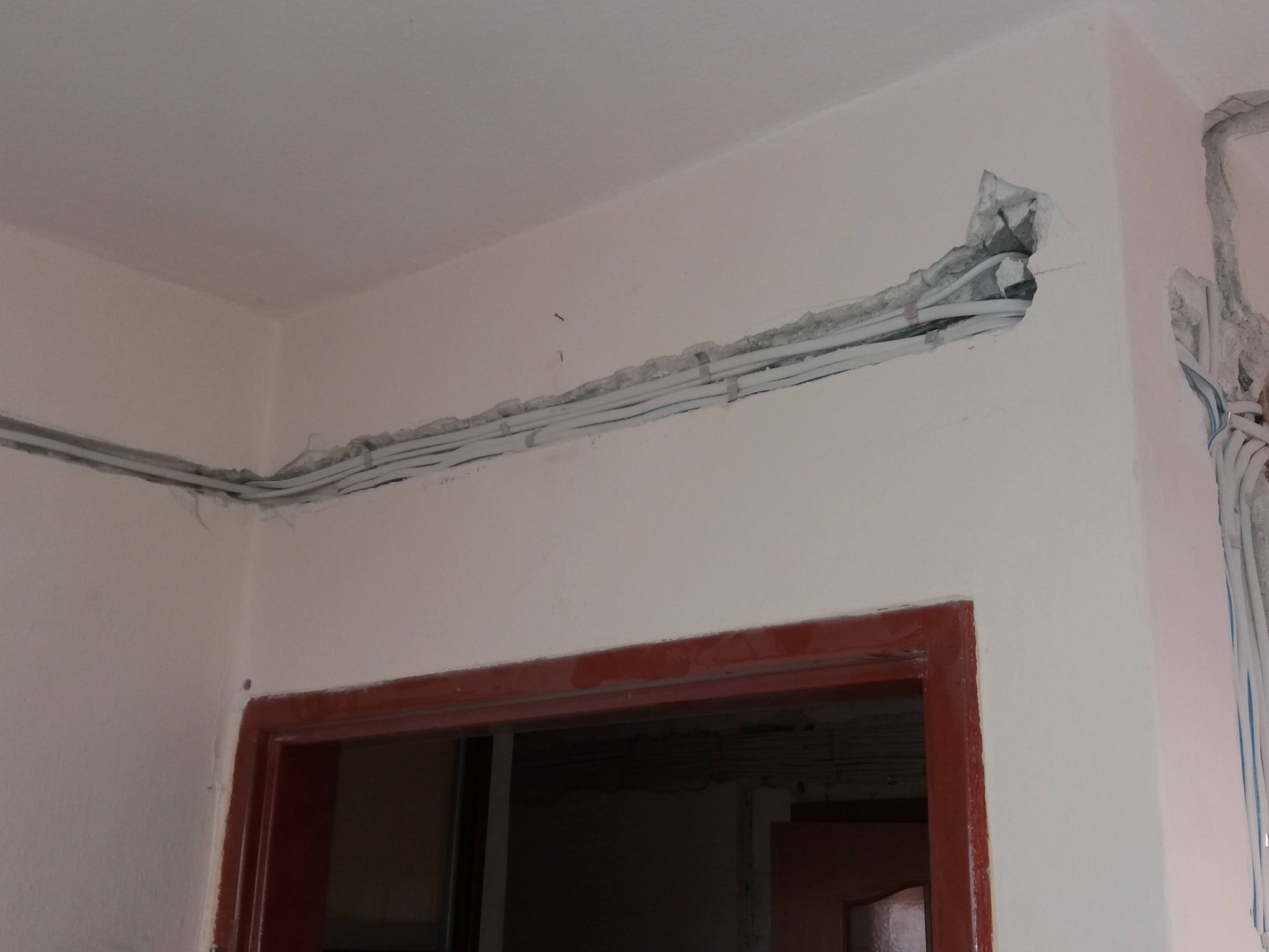Białe przewody elektryczne ułożone w bruździe w ścianie, widoczny fragment instalacji w trakcie prac wykończeniowych, surowy stan ściany z widoczną strukturą betonu.