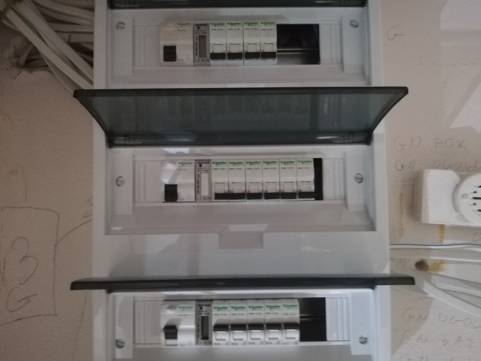 Rozdzielnica elektryczna z częściowo otwartą klapką, widoczne bezpieczniki i kable, ściana z notatkami ołówkiem w tle.