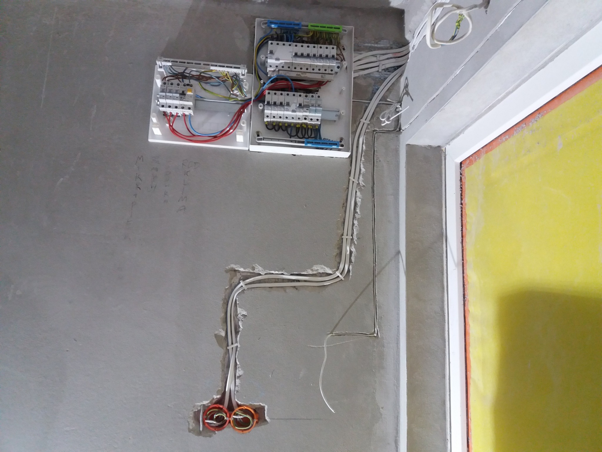 Rozdzielnice elektryczne z okablowaniem na surowej ścianie, widoczne przewody w peszlach i puszki instalacyjne, ściana z notatkami ołówkiem.