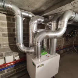 Centrala rekuperacyjna WANAS z podłączonymi przewodami wentylacyjnymi w surowym pomieszczeniu z ceglanymi ścianami i betonowym sufitem.