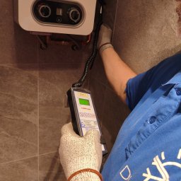 Duoterm - Adam Kurasz - Sprawdzanie szczelności pieca gazowego detektorem EDG-1. Mężczyzna w rękawicach ochronnych przy piecu na tle szarej ściany. Widoczny fragment niebieskiej koszulki.
