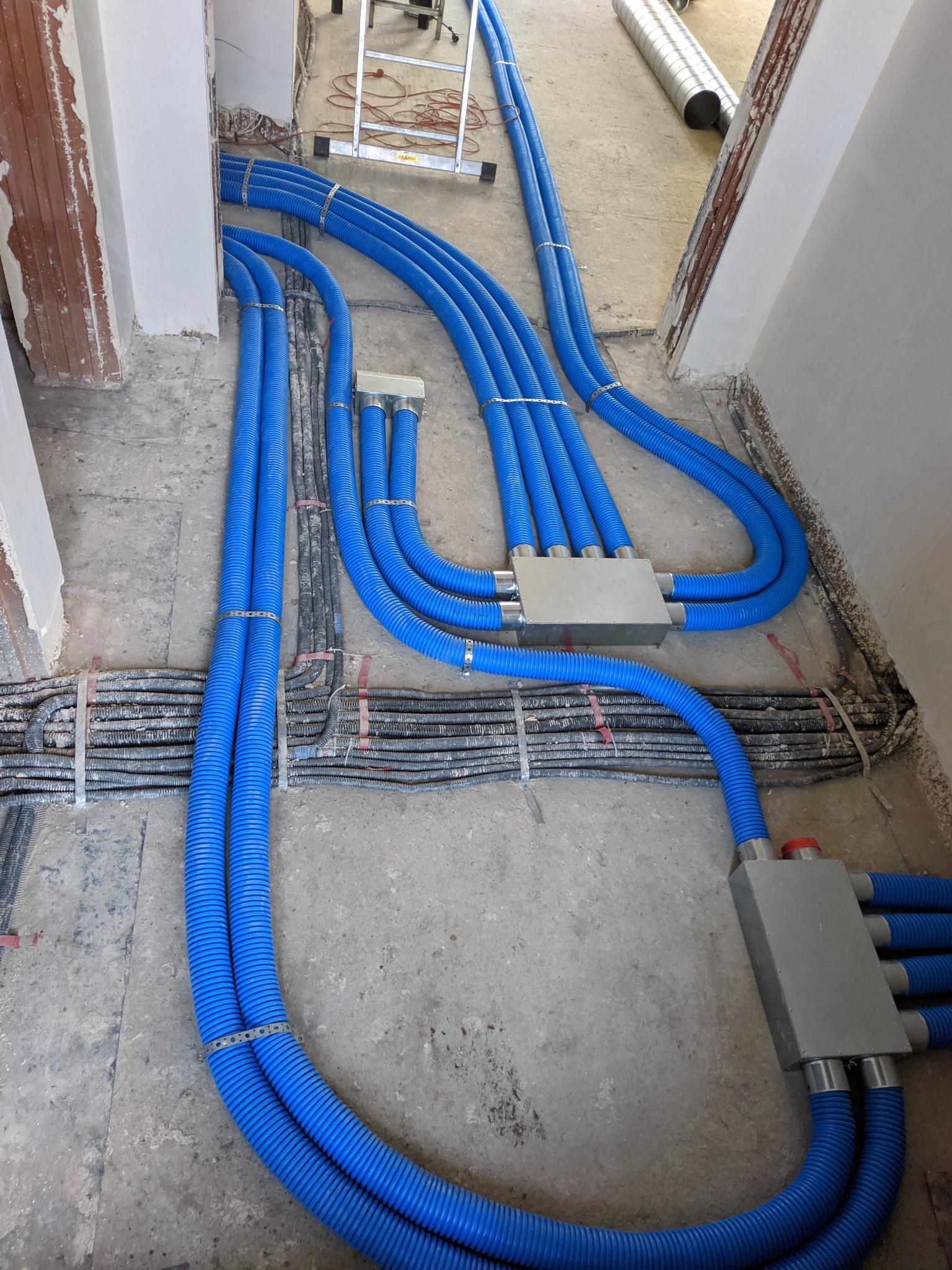 Niebieskie rury wentylacyjne ułożone na podłodze w trakcie montażu, połączone metalowymi skrzynkami rozdzielczymi, widoczny fragment drabiny i rura wentylacyjna w tle.