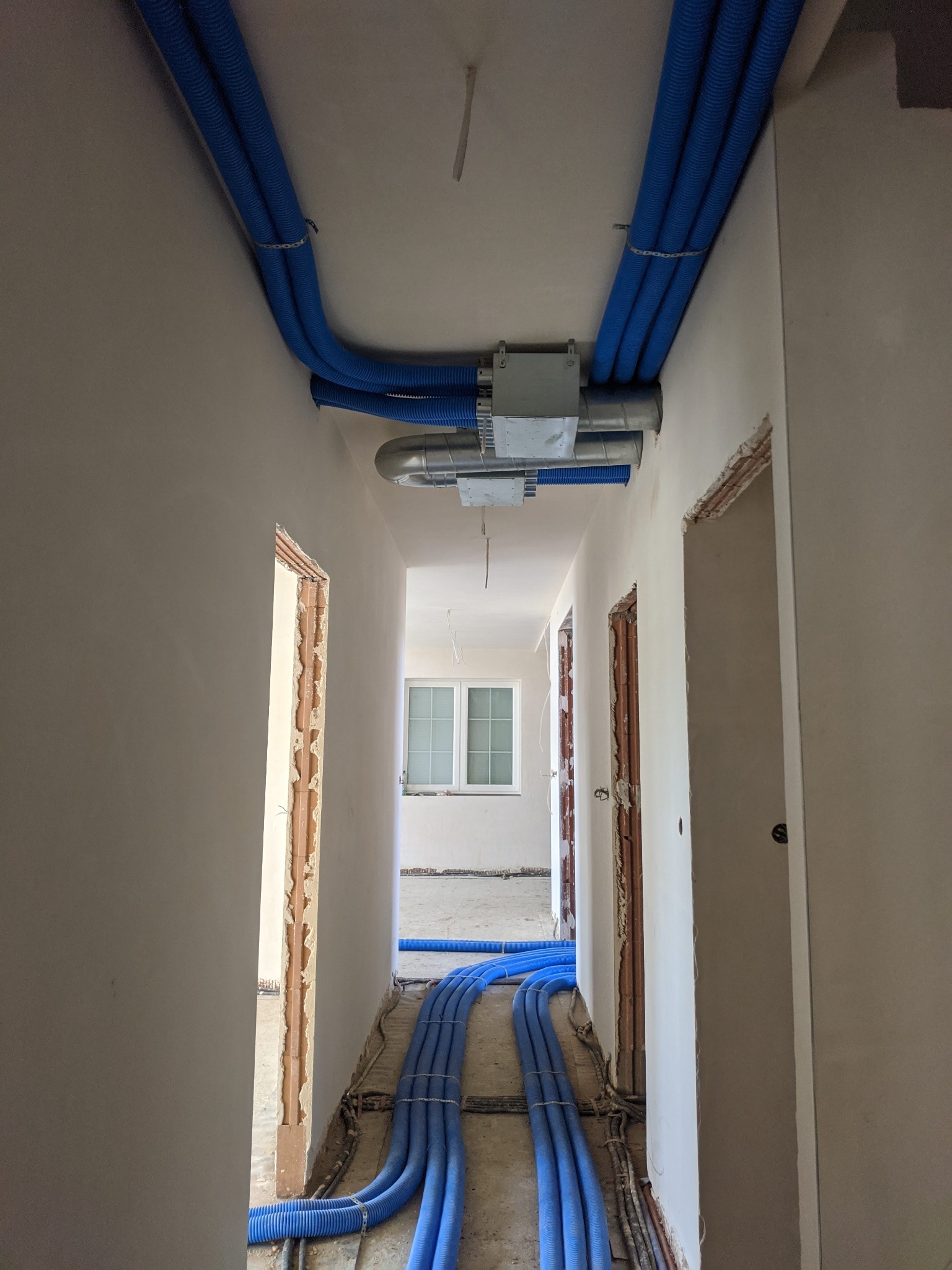 Wnętrze w trakcie prac wykończeniowych z widocznymi niebieskimi rurami wentylacyjnymi poprowadzonymi pod sufitem i na podłodze, centrala wentylacyjna zamontowana na suficie, surowe ościeżnice...