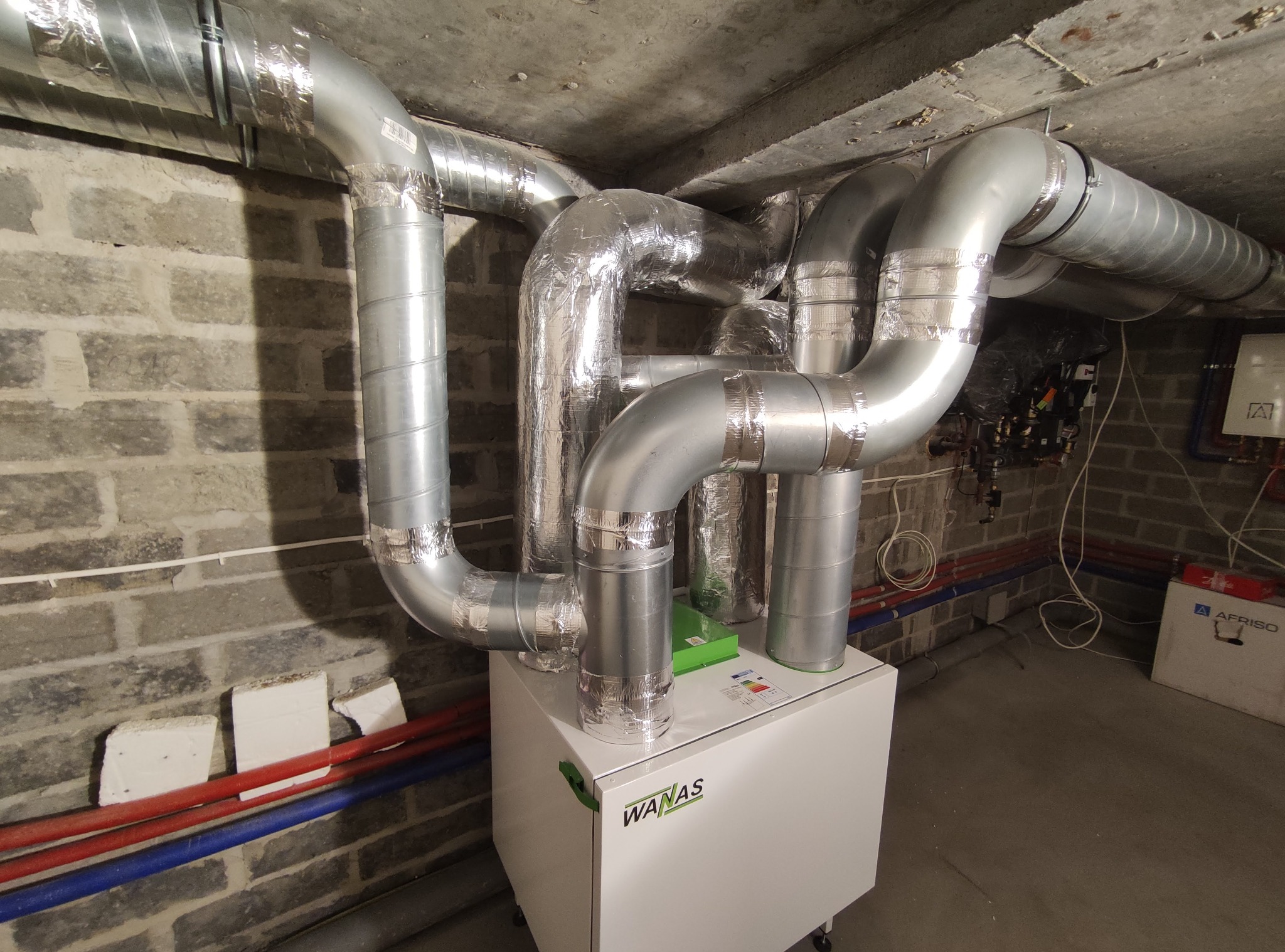 Centrala rekuperacyjna WANAS z podłączonymi przewodami wentylacyjnymi w surowym pomieszczeniu z ceglanymi ścianami i betonowym sufitem.