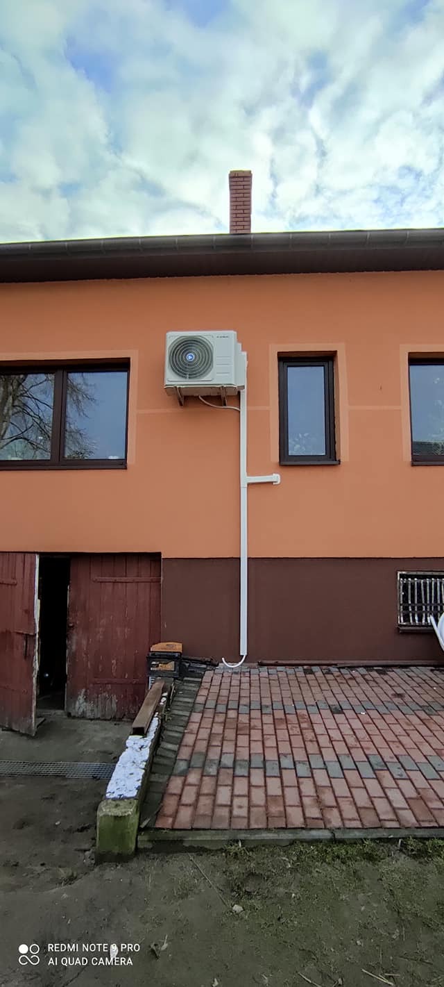 Zewnętrzna jednostka klimatyzacji zamontowana na pomarańczowej ścianie budynku, z widocznymi rurami odprowadzającymi skropliny. Na pierwszym planie brukowany chodnik i otwarte drzwi do garażu.