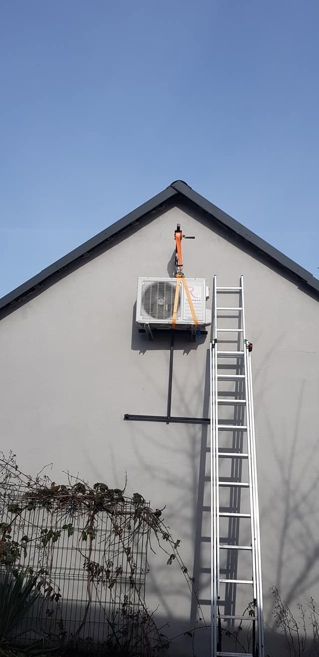 Montaż jednostki zewnętrznej klimatyzatora na ścianie budynku, zabezpieczonej pasami transportowymi, z wykorzystaniem drabiny aluminiowej.