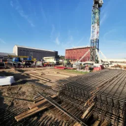 Siatka zbrojeniowa na budowie z widocznym dźwigiem, szalunkiem i budynkiem w tle, w słoneczny dzień.