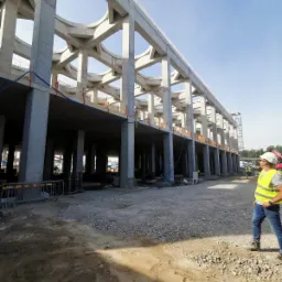 Surowa konstrukcja żelbetowa budynku w trakcie budowy, widok z dołu, pracownik w kamizelce odblaskowej na pierwszym planie, widoczne zbrojenia i szalunki.