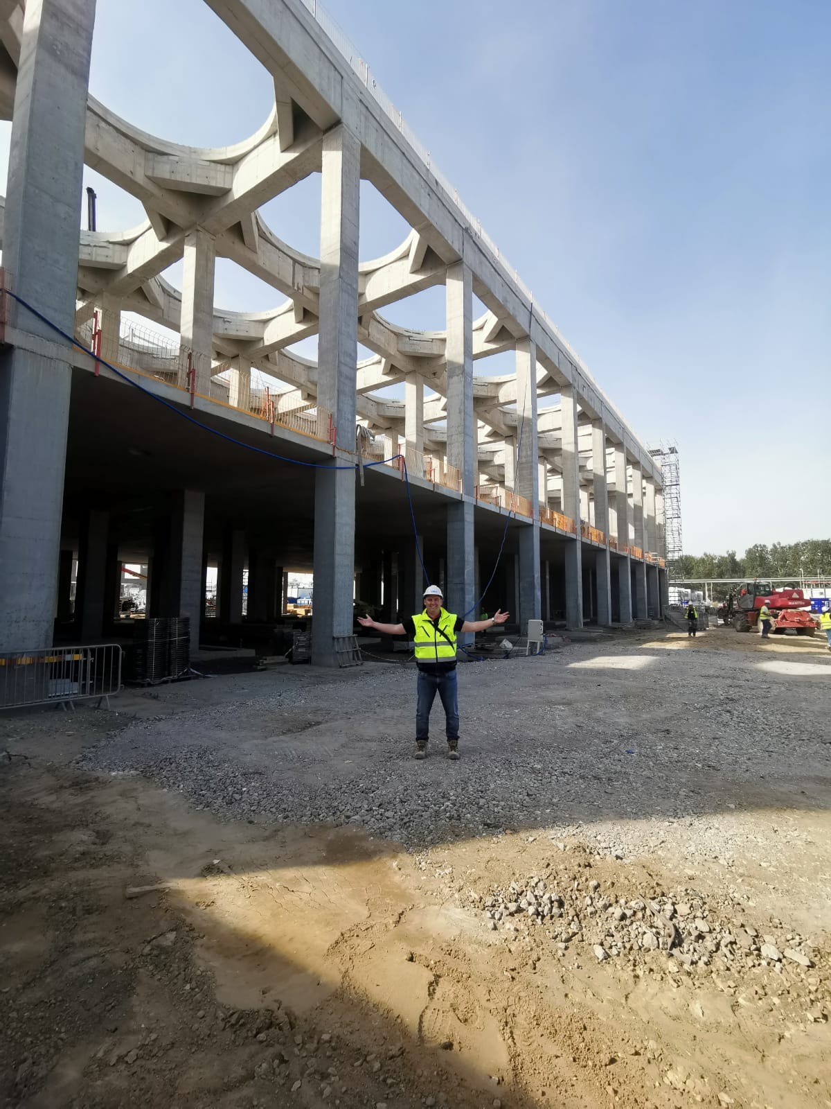 Konstrukcja żelbetowa w trakcie budowy, widok z dołu na szkielet budynku z kolumnami i charakterystycznymi łukowymi połączeniami, na pierwszym planie pracownik w kasku i kamizelce odblaskowej...