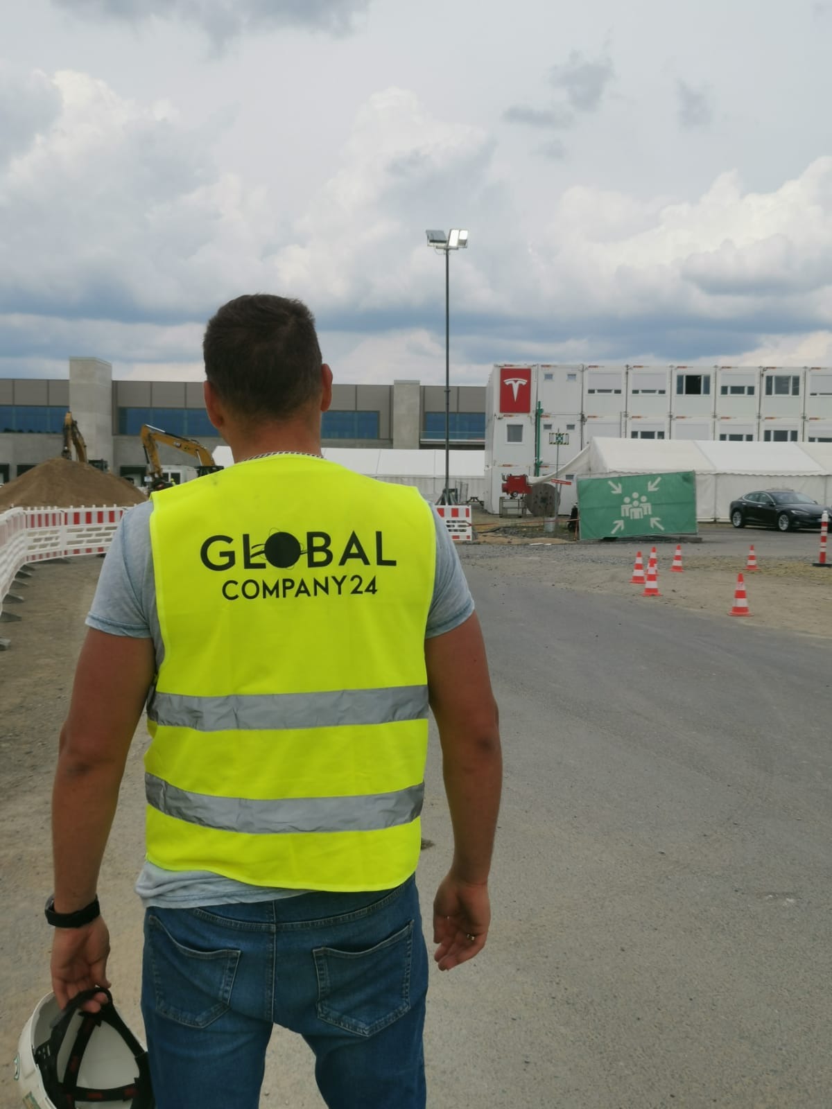 Pracownik w żółtej kamizelce z logo firmy budowlanej na tle budowy, widoczny dźwig i baraki.