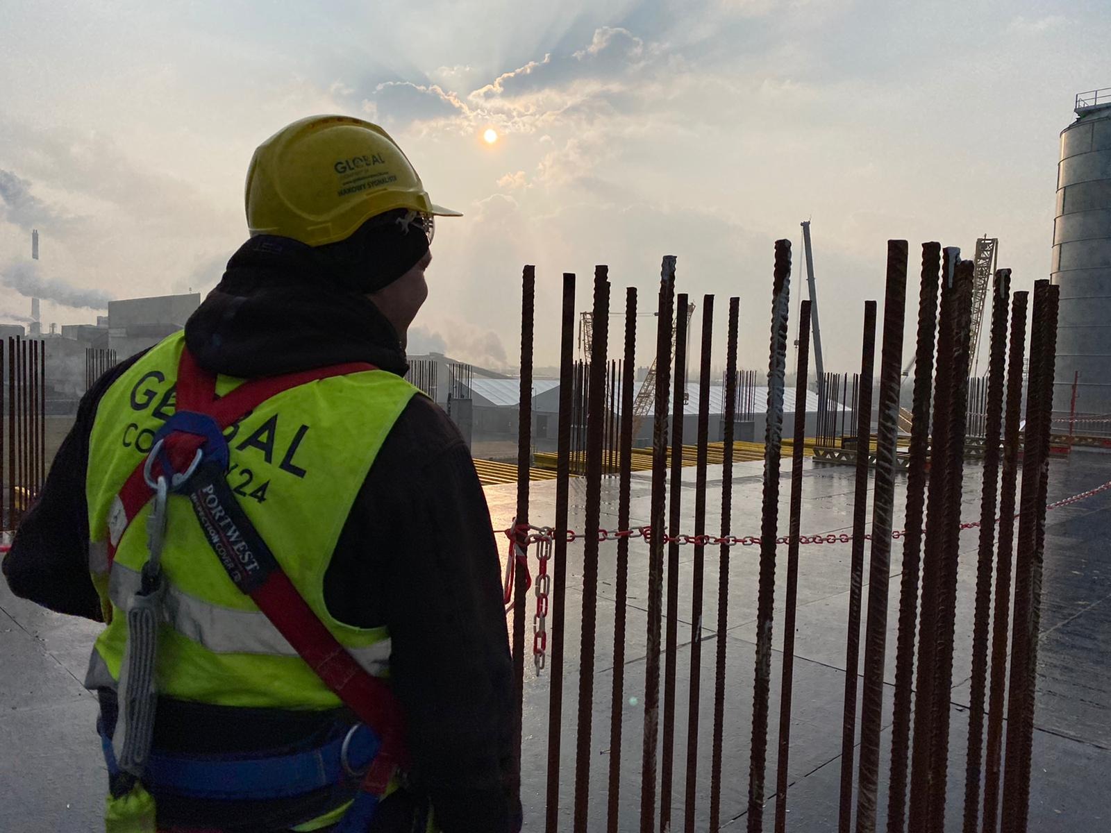 Pracownik w żółtym kasku i kamizelce odblaskowej, zabezpieczony szelkami, stoi na tle zbrojenia pionowego i rozległego placu budowy z widocznym dźwigiem i kominami fabrycznymi w oddali...