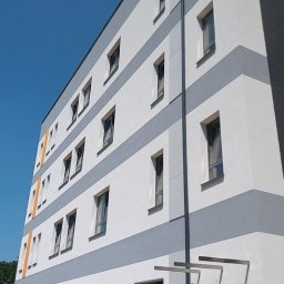 LIDER-BUD Sp. z.o.o - Wielopiętrowy budynek z nową, biało-szarą elewacją, z widocznymi oknami i rurą spustową na tle błękitnego nieba.