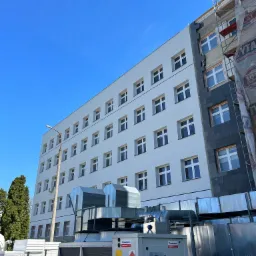 Wielopiętrowy budynek częściowo ocieplony styropianem, z widocznymi rusztowaniami i instalacją wentylacyjną na zewnątrz, na tle błękitnego nieba.