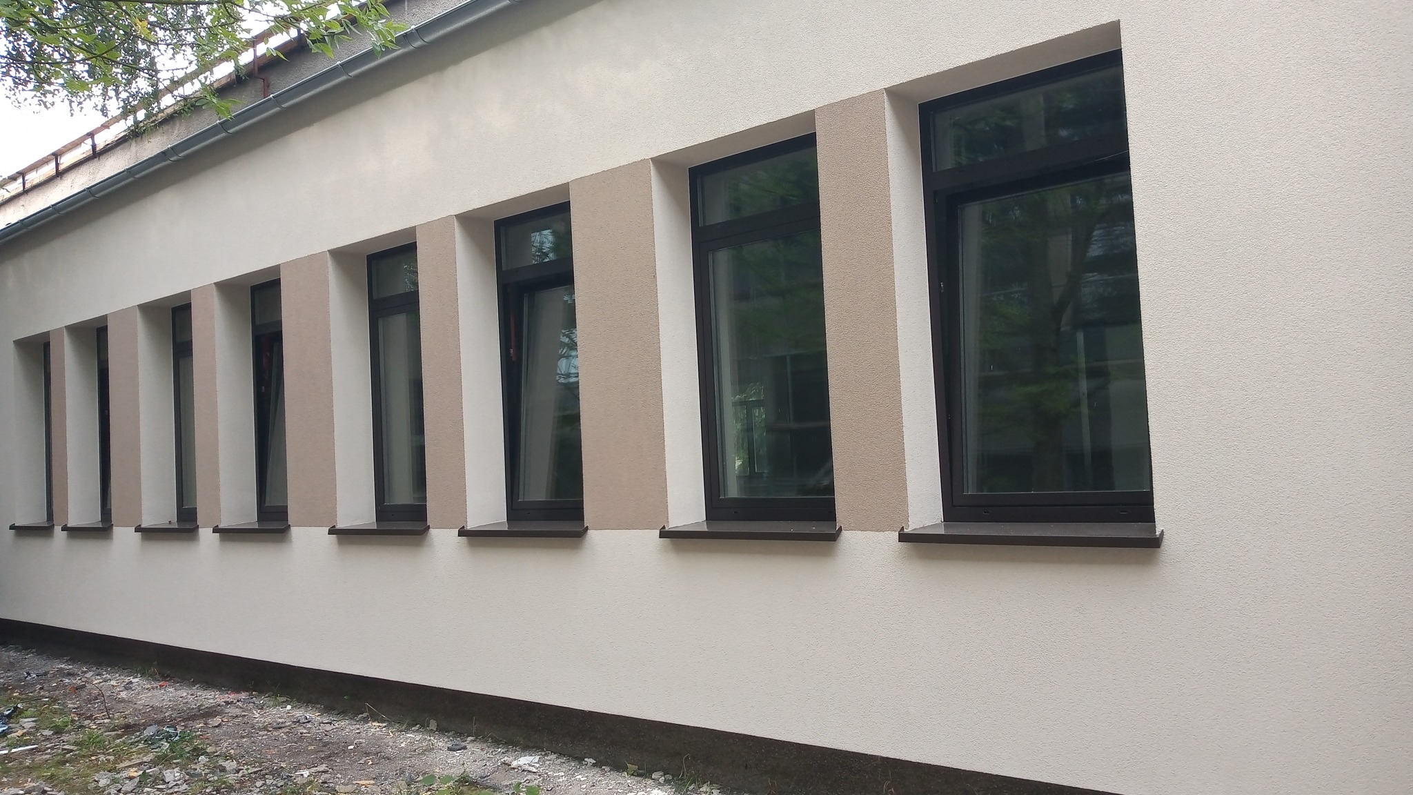 Odnowiona elewacja budynku z szeregiem okien w ciemnych ramach, wykończona tynkiem w odcieniach beżu i bieli, z widocznymi śladami prac ziemnych u podstawy.