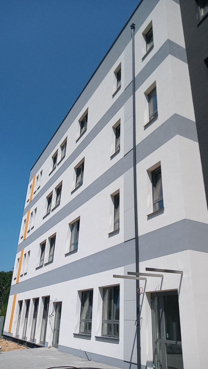 Wielopiętrowy budynek z nową, biało-szarą elewacją, z widocznymi oknami i rurą spustową na tle błękitnego nieba.