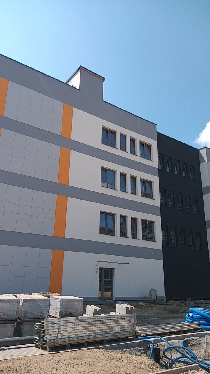 Nowoczesny budynek wielorodzinny z elewacją w odcieniach szarości, bieli i pomarańczy, widoczne materiały budowlane na pierwszym planie, sugerujące prace wykończeniowe.