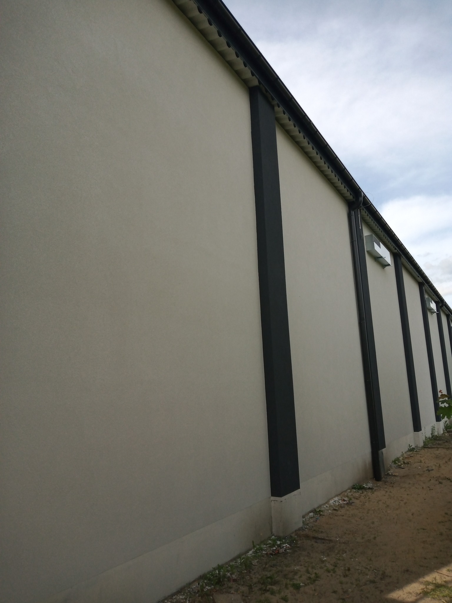 Jasnoszara elewacja budynku z ciemnymi, pionowymi elementami konstrukcyjnymi, widok z dołu, z widocznym fragmentem rynny i terenu przy budynku.