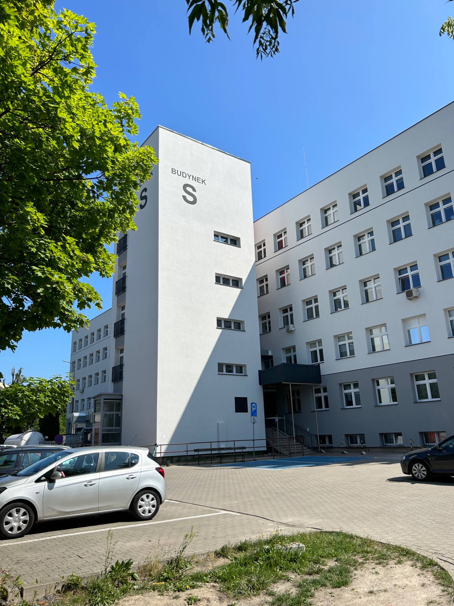 Wysoki, biały budynek z napisem 'BUDYNEK S' i dużymi literami 'S', widoczny z perspektywy parkingu w słoneczny dzień.