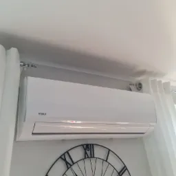 Biały klimatyzator marki Vivax zamontowany na ścianie w jasnym pomieszczeniu, obok dekoracyjny zegar w stylu industrialnym i białe zasłony.