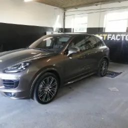 Brązowe Porsche Cayenne 4S na stanowisku warsztatowym z napisem 'POSTFACTORY.PL' w tle, widoczne felgi i reflektory.
