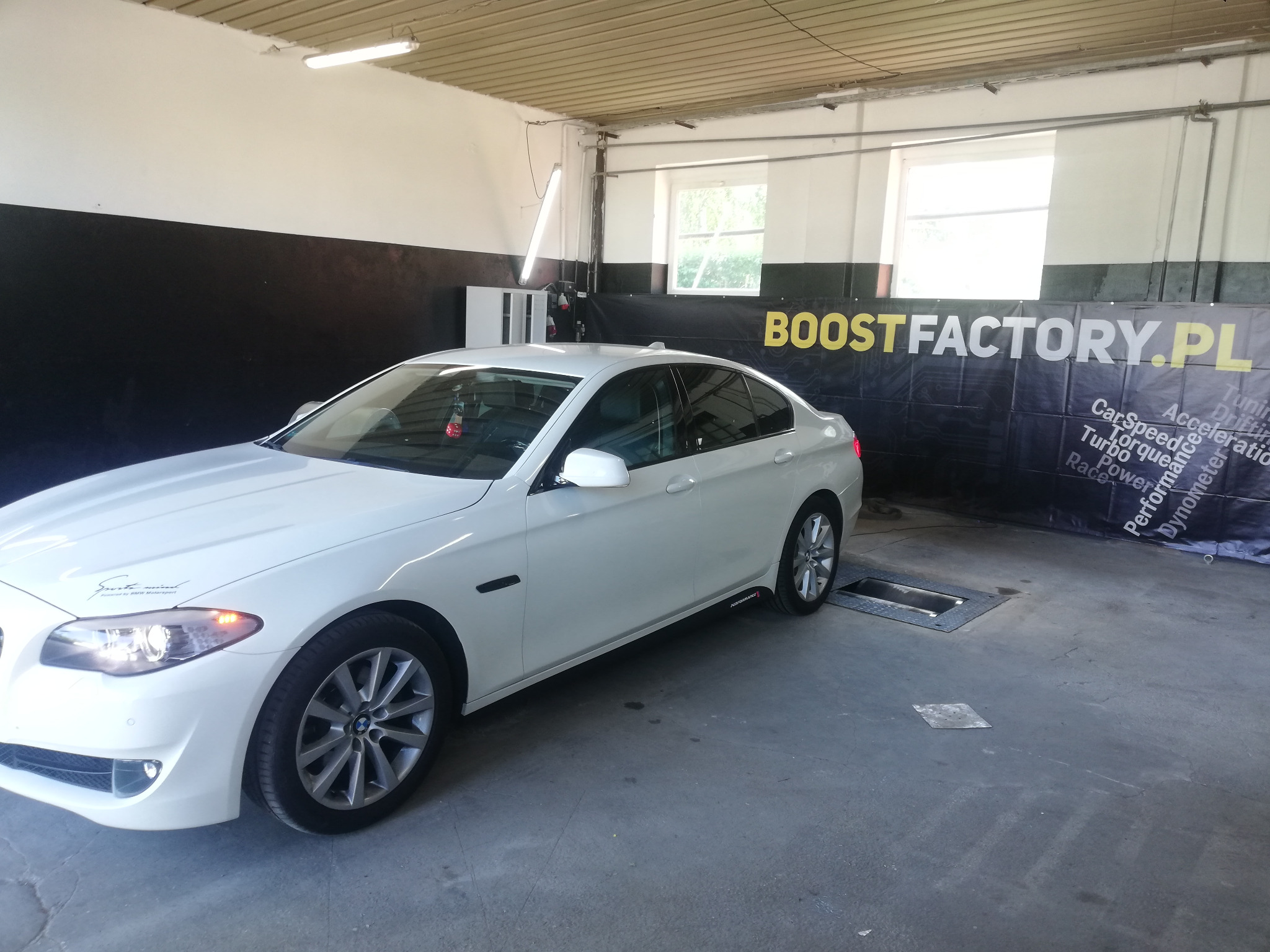 Białe BMW F10 na stanowisku diagnostycznym w warsztacie, widoczny baner Boostfactory.pl w tle.