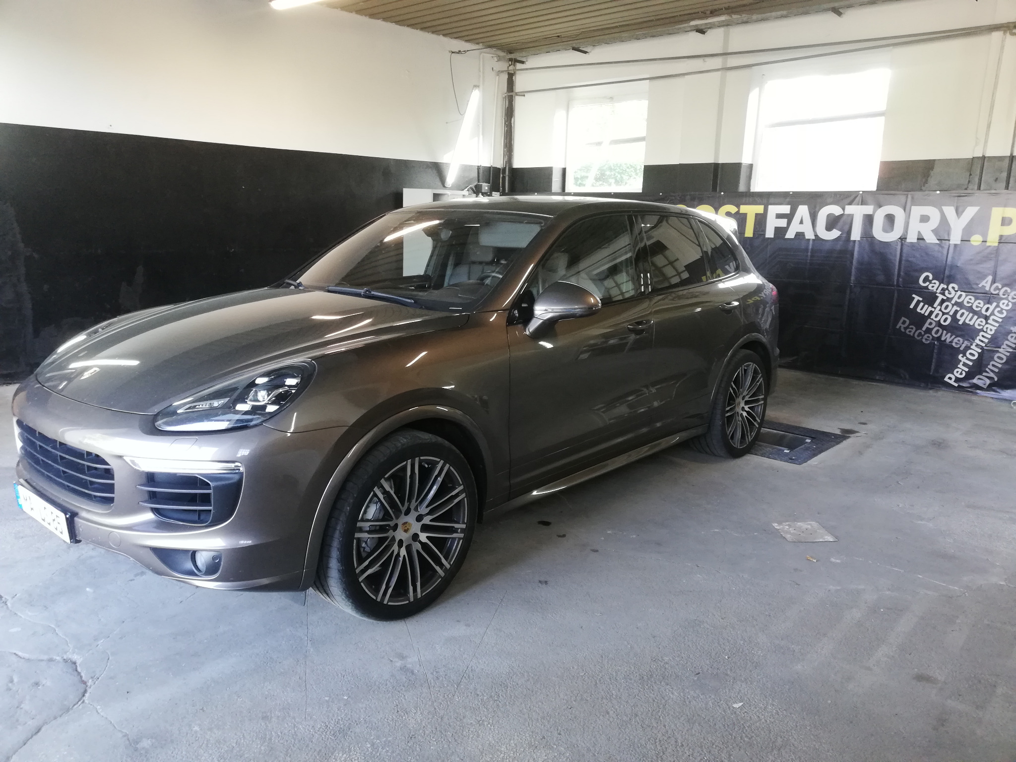Brązowe Porsche Cayenne 4S na stanowisku warsztatowym z napisem 'POSTFACTORY.PL' w tle, widoczne felgi i reflektory.