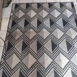 Dywan z geometrycznym wzorem w odcieniach szarości, częściowo zwinięty, obok wiertarka z żółtą szczotką do czyszczenia, leżący na kafelkach podłogowych.