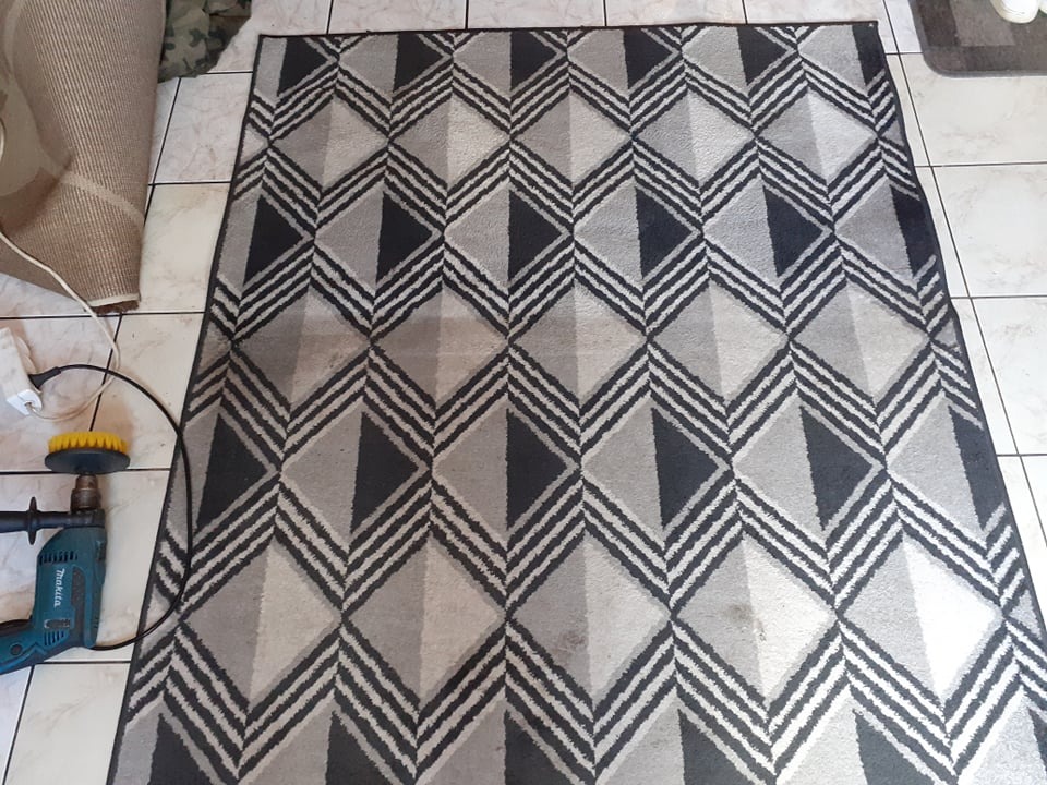 Dywan z geometrycznym wzorem w odcieniach szarości, częściowo zwinięty, obok wiertarka z żółtą szczotką do czyszczenia, leżący na kafelkach podłogowych.