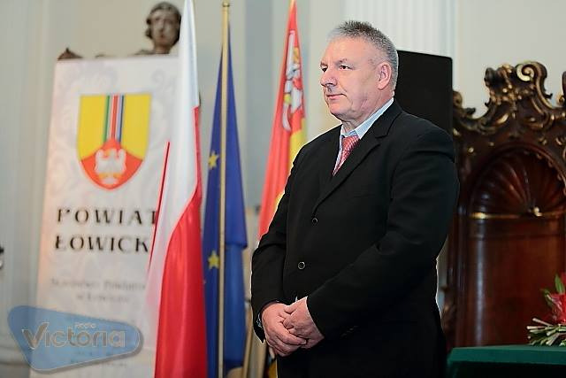 Uroczyste wystąpienie mężczyzny w garniturze na tle flagi Polski, flagi Unii Europejskiej i herbu Powiatu Łowickiego.