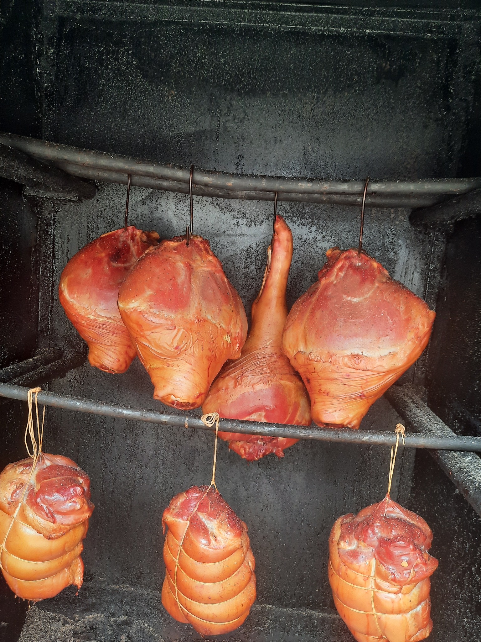 Wędzone szynki i balerony wiszące na metalowych prętach w wędzarni z ciemnym, osmolonym wnętrzem.