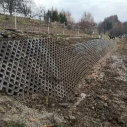 Wzmocnienie skarpy z użyciem ażurowych płyt betonowych, widoczne ślady prac ziemnych i błoto, w tle drzewa i pochmurne niebo.