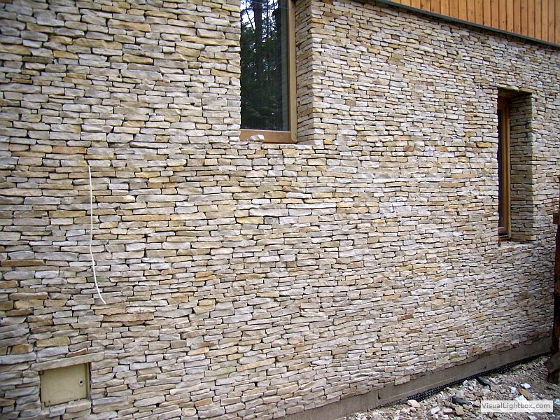 Elewacja budynku wykończona nieregularnym kamieniem piaskowcem w odcieniach beżu i szarości, widoczne dwa okna z drewnianą ramą i wystający kabel.
