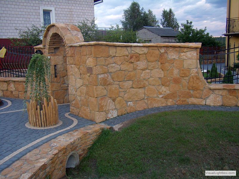 Ogród z elementami z piaskowca: murki, łuk, obudowa rośliny i obrzeża, w tle dom i trawnik.