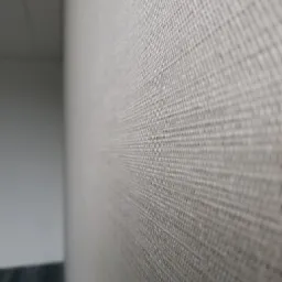 Delikatna, beżowa tapeta o wyraźnej strukturze tkaniny, przylegająca do ściany, z widocznym fragmentem szarej podłogi w dolnym rogu kadru i białym sufitem w górnym rogu.