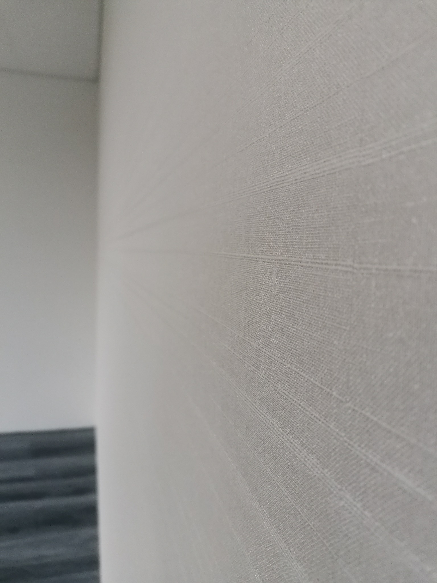 Ściana pokryta jasną tapetą o delikatnej fakturze w Gliwicach, widoczny fragment podłogi z szarymi panelami.