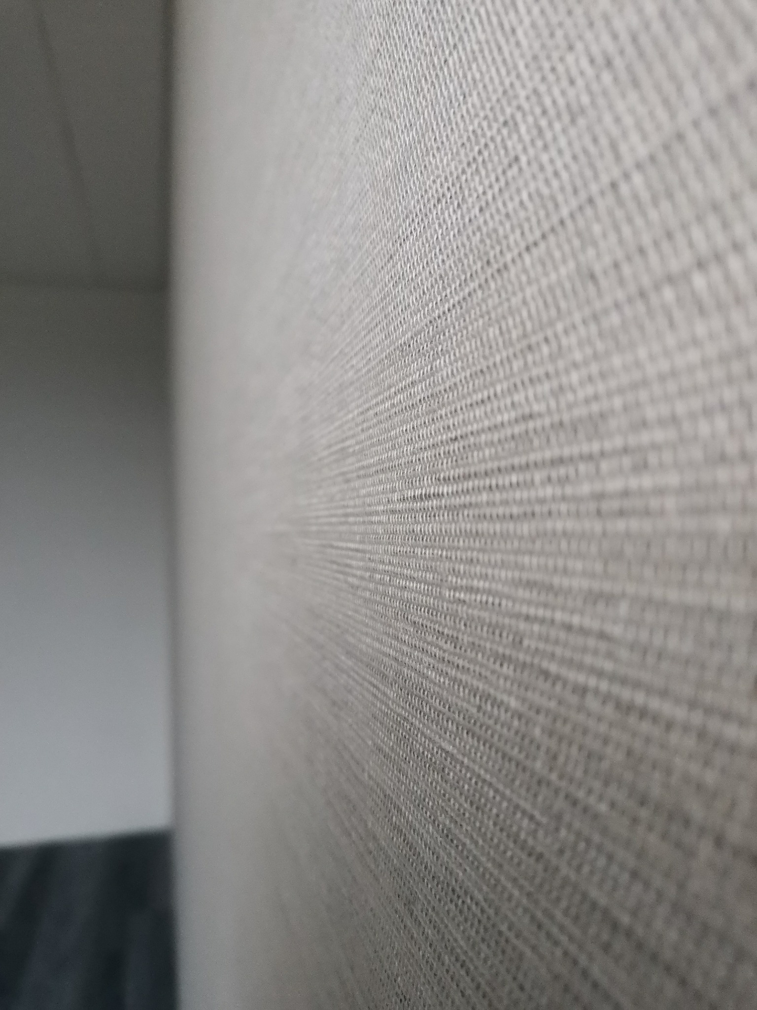 Delikatna, beżowa tapeta o wyraźnej strukturze tkaniny, przylegająca do ściany, z widocznym fragmentem szarej podłogi w dolnym rogu kadru i białym sufitem w górnym rogu.