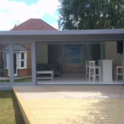 Nowoczesna altana z drewnianym tarasem, przeszklonymi ścianami i rozsuwanymi drzwiami, wnętrze z barem i fototapetą z motywem plaży.