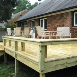 Nowo wybudowany taras z jasnego drewna z barierkami, schodami i zestawem mebli ogrodowych na tle ceglanego domu.