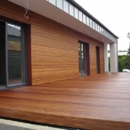 Nowoczesny dom z elewacją z jasnego drewna i przesuwnymi drzwiami, z przylegającym tarasem z egzotycznego drewna o ciepłym, brązowym odcieniu.